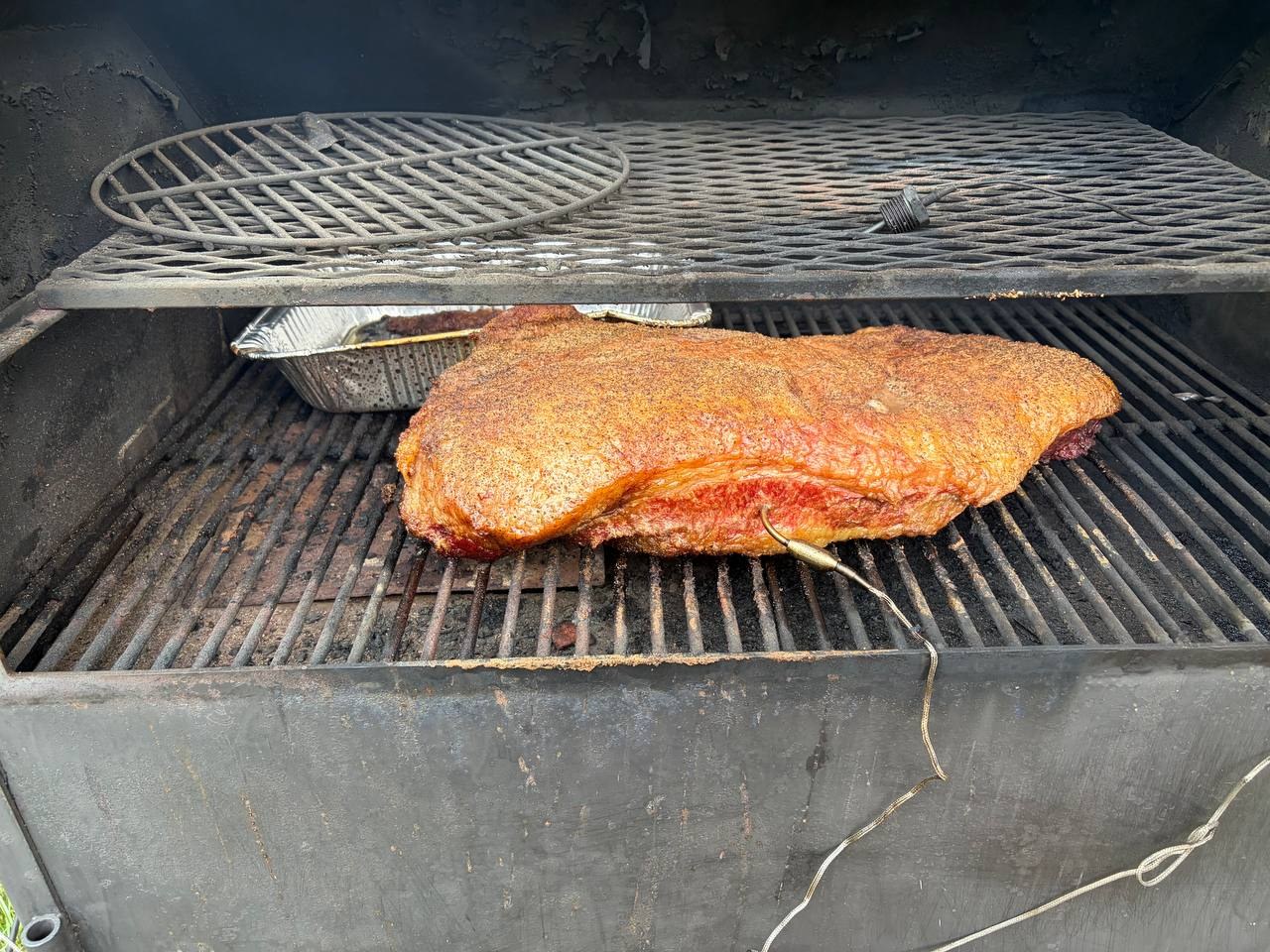 A brisket cooking unwrapped on the lower smoker grate, with a temperature probe in the side and bark beginning to form on the glossy red-orange surface.