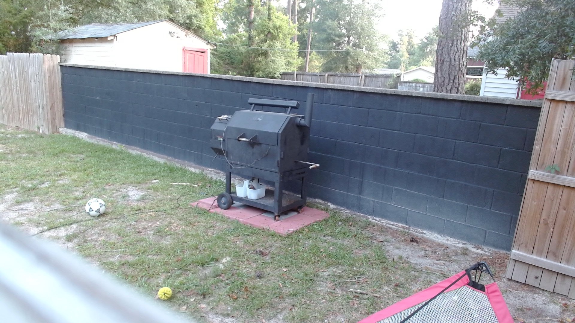Morning webcam view of the backyard smoker closed against a black wall, with probe wiring visible and no obvious smoke or flame issue.