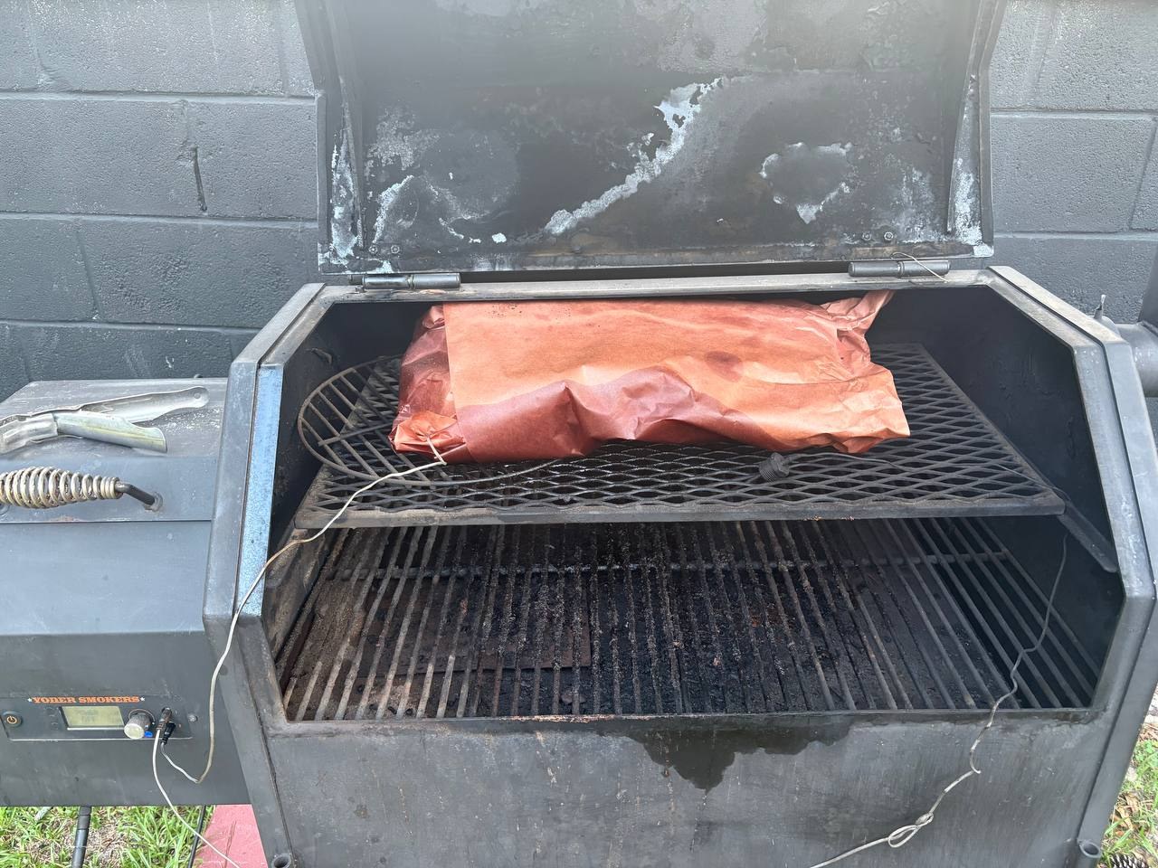 A butcher-paper-wrapped brisket resting on the top rack of an open Yoder smoker, with probe wires visible across the grate.