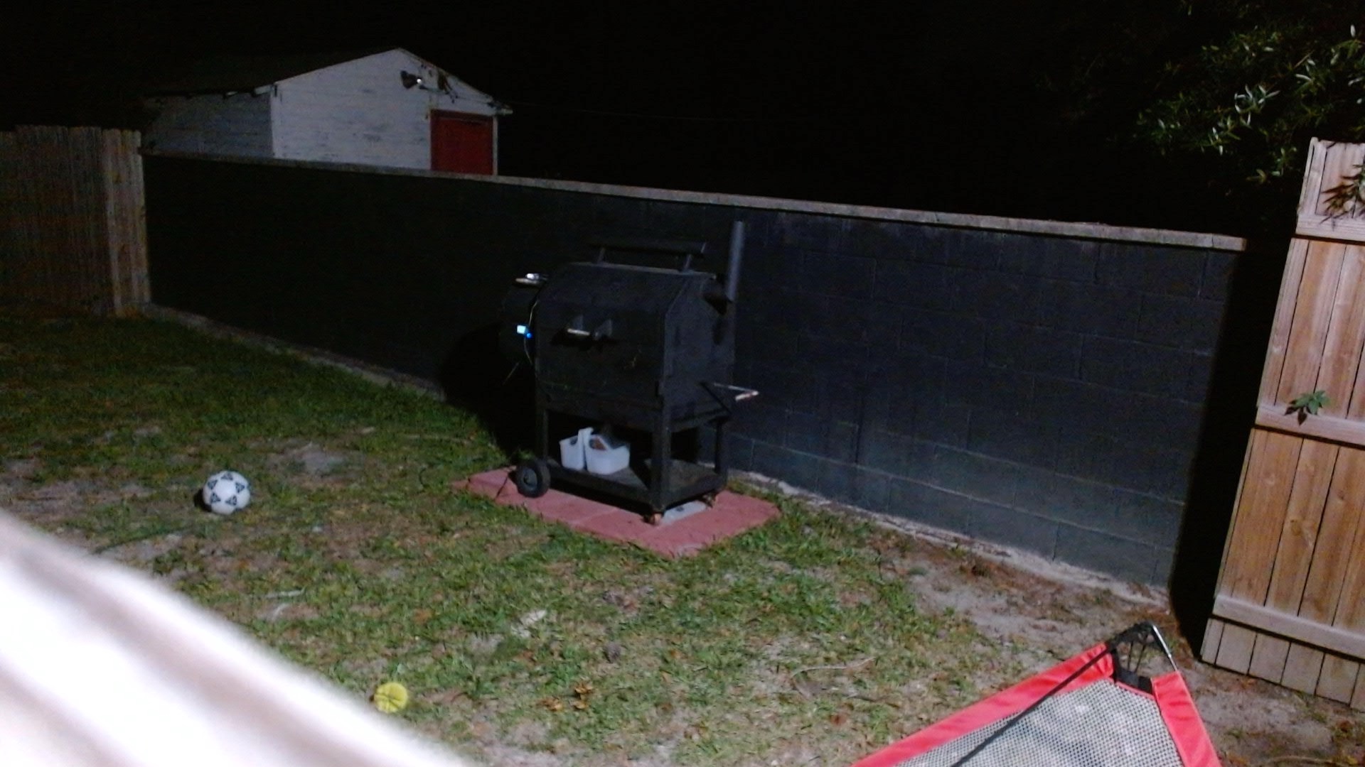 A nighttime webcam view of the backyard smoker closed against a black wall, with the FireBoard light visible and no obvious smoke or flame issue.