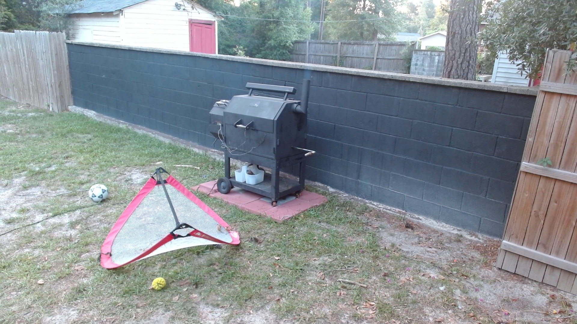 A backyard webcam view of the closed offset smoker against a black wall, with FireBoard probe wires visible and evening light in the yard.
