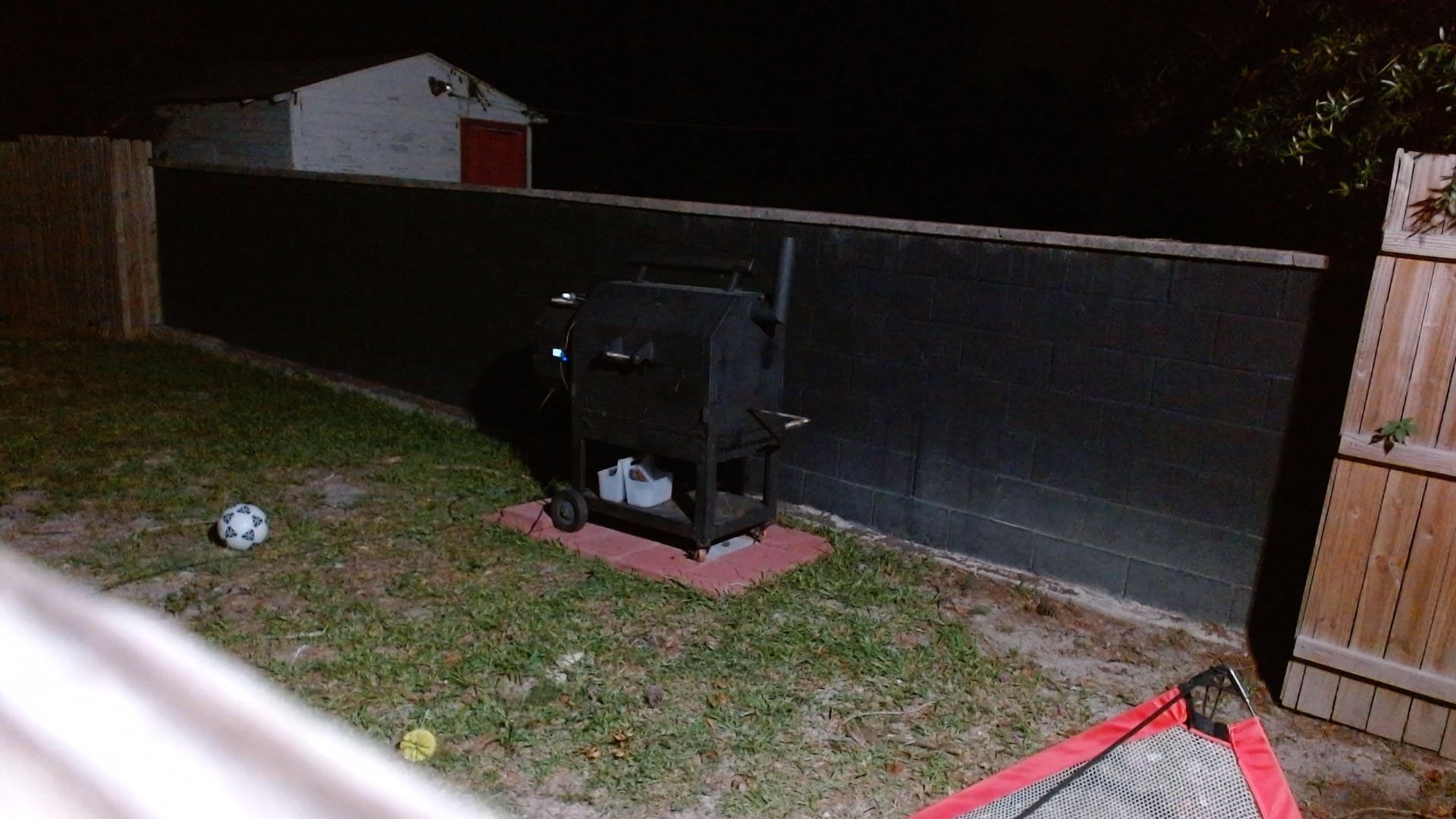 A nighttime backyard webcam view of the closed offset smoker against a black wall, with FireBoard wiring visible and no obvious smoke or fire issue.