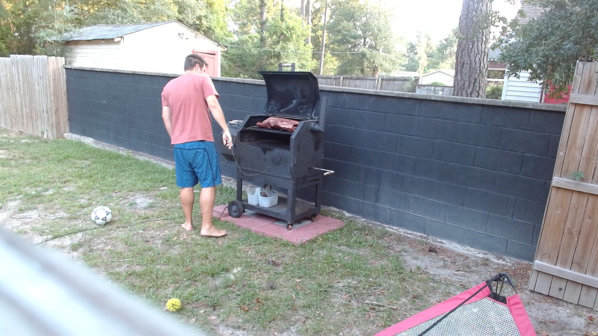 Morning webcam view of Justin tending the open backyard smoker, with the wrapped brisket visible on the grate and no obvious smoke or flame issue.